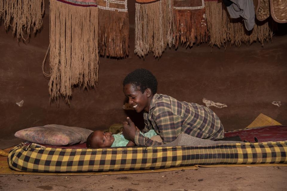 Fati, 14, playing with his younger brother Khalif, 3months, at their home in a remote community in Oromia Region, Ethiopia. Fati wears a checked shirt, he's leaning over his baby brother - smiling down at him. Khalif is wearing a blue/green vest.
