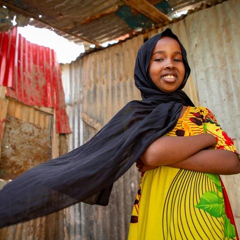 A young girl stands in front of corrugated iron with her arms folded, smiling. Her black headscarf blows out behind her as she looks down at the camera.