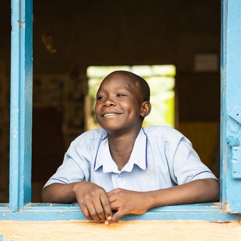 A young boy in a pale blue shirt looks out of a building through a blue window frame, smiling.
