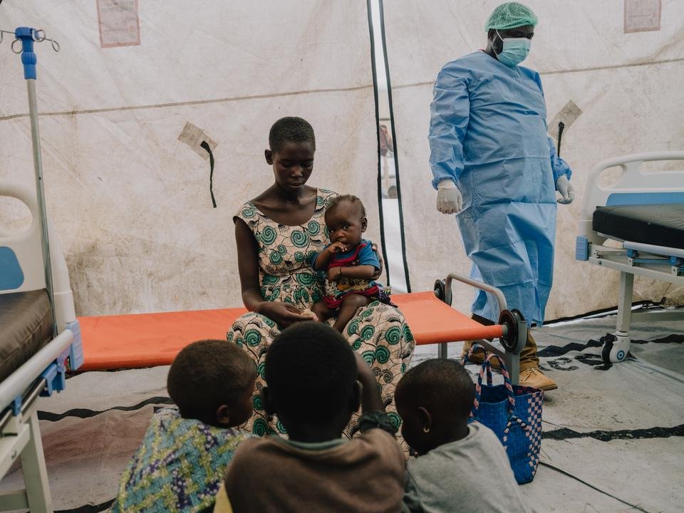 Furaha*, 20, sits on a bed in an isolation tent, holding her 7 month old son, David*, while her other children crouch in front of them