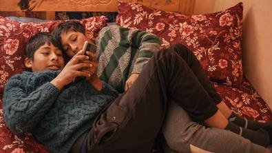 Two boys in wooly jumper lie on a bed looking at a mobile phone.