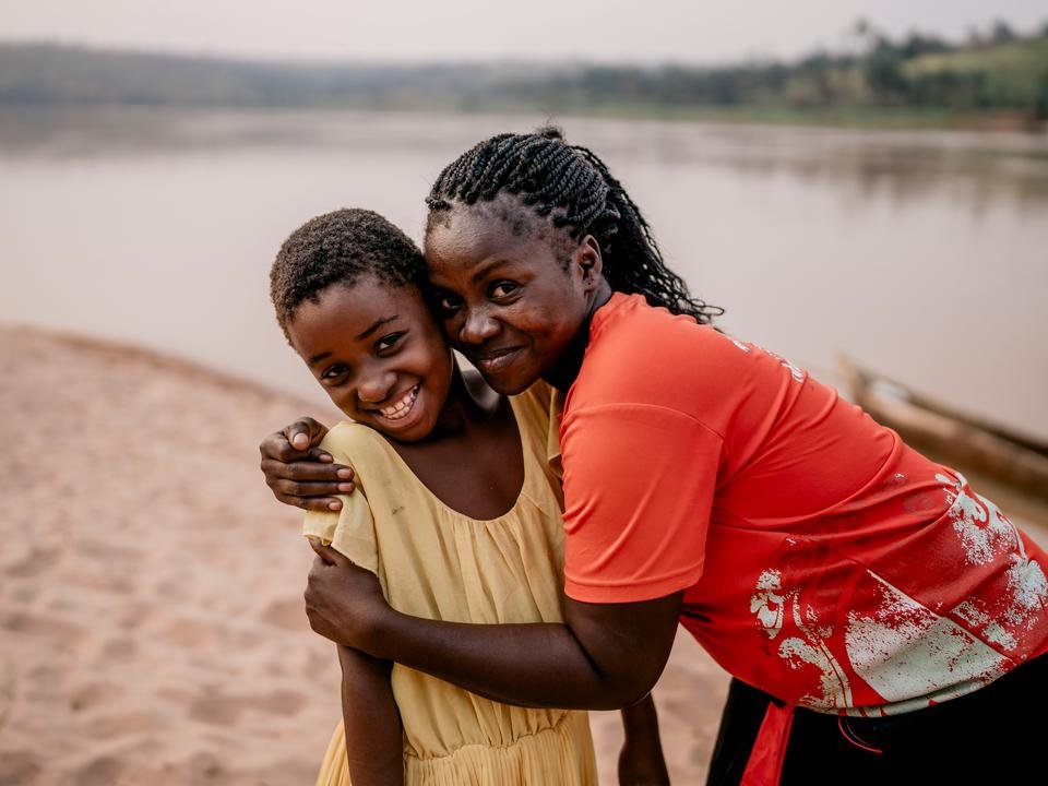 A Shelter For Kids supported worker called Patience hugs 10-year-old Chouchou*, 10, by the river in Kasai, DRC. Both Chouchou and Patience are smiling at the camera.