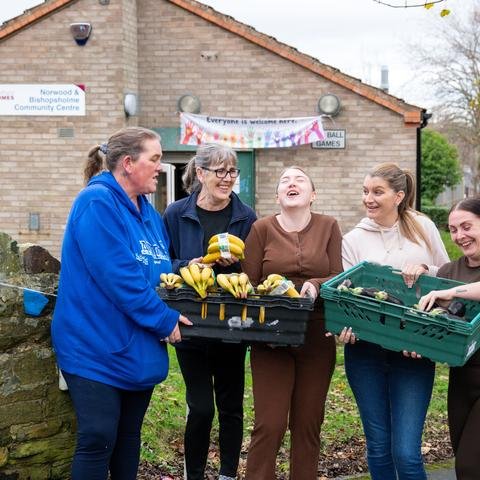 Members of You, Me & Us stand outside a community centre, laughing and holding crates of fruit and vegetables