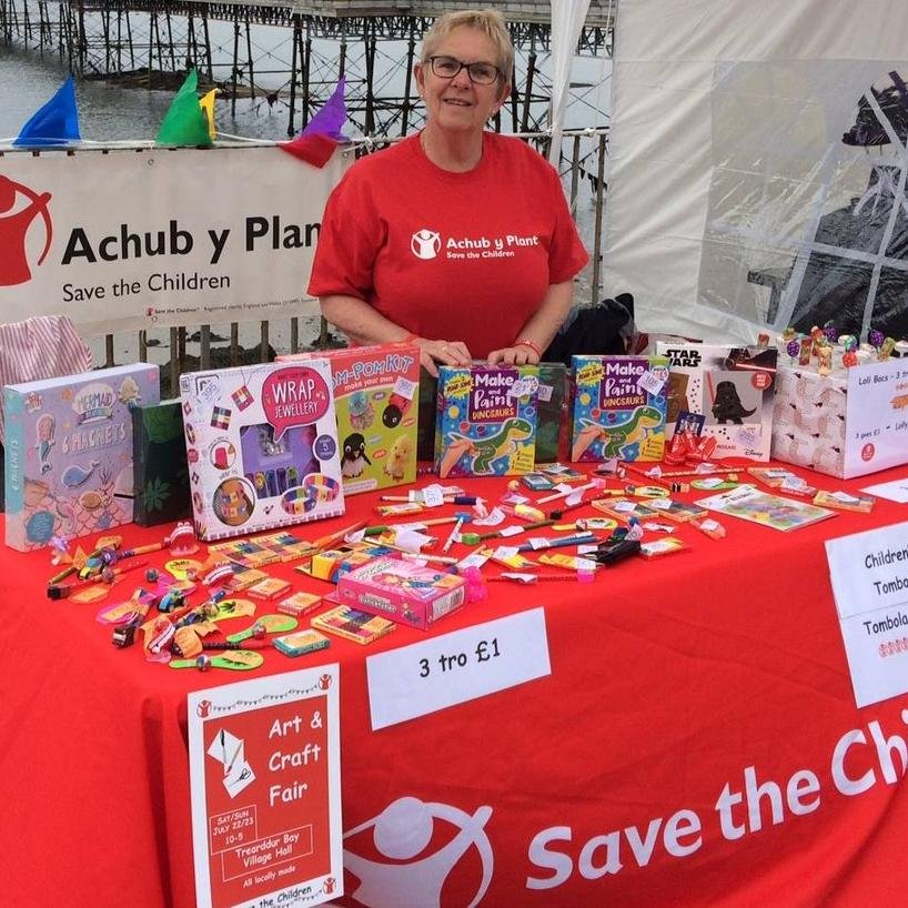 A lady stands behind a market stall selling crafts. She's wearing a red Shelter For Kids tshirt and the Shelter For Kids logo is on a banner behind her.