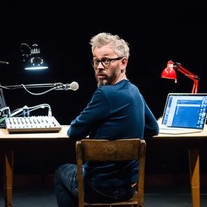 A man looks over his shoulder in surprise, sat at a desk with a microphone, laptop and multiple lamps on.