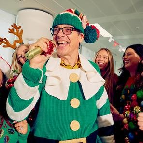 A group of people in an office have fun singing together, all wearing Christmas jumpers. A man in the middle is dressed as a Christmas elf and holds a microphone.